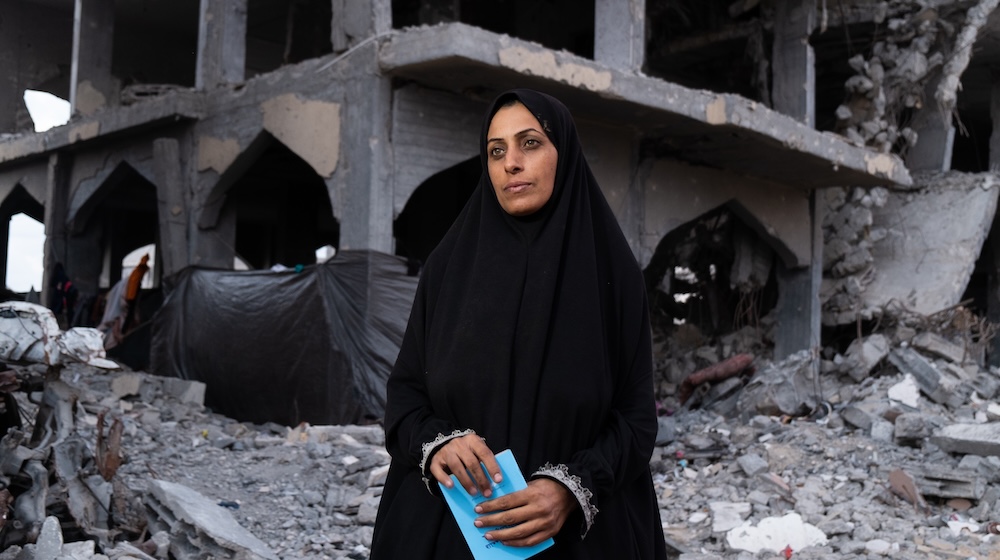 A woman wearing a black chador stands in front of the rubble of a destroyed building.