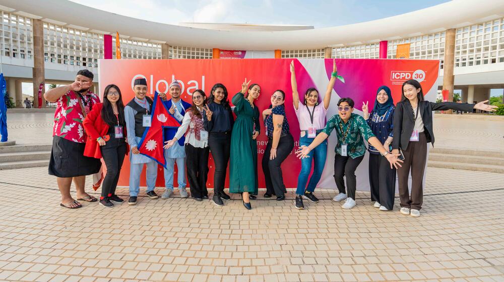 A group of young students in blue uniforms raise their hands while seated in a classroom