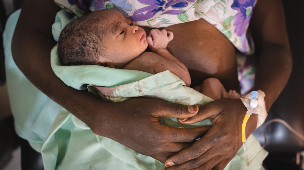 An adolescent girl holds her newborn daughter in a bloodied blanket. There is an IV line in the girl’s wrist.