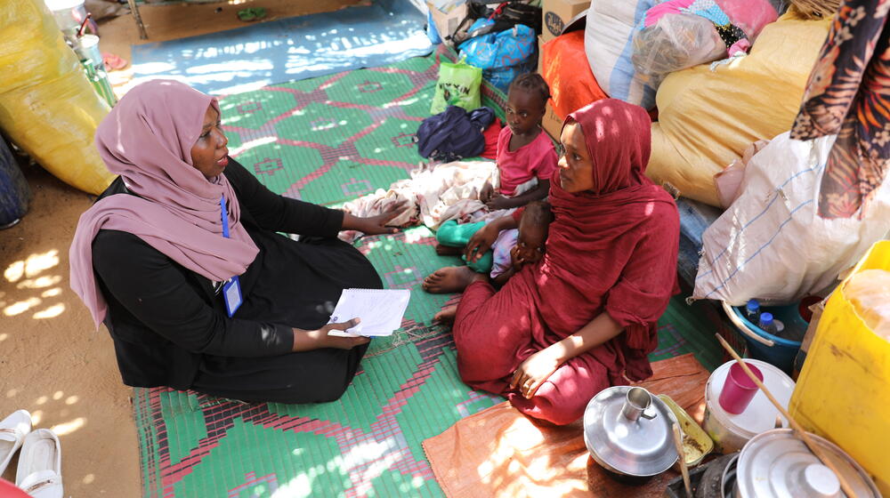 A woman in red sits beside two of her children on a colorful blanket over sand. She speaks to a woman in a headscarf and badge.
