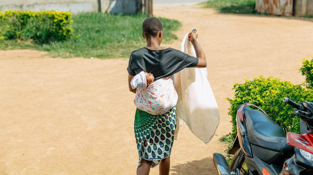 A young woman in a dark top and colourful patterned skirt walks away with a young baby strapped to her back and carrying a large white sack
