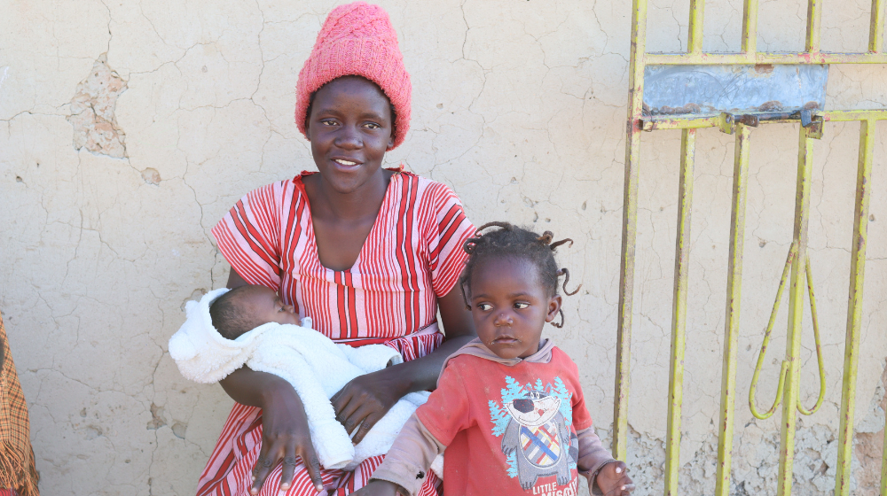 A woman in a dress and a pink hat holds a newborn in a white fleece. A young child stands beside her.