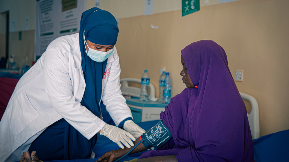  A woman in a white coat, blue head scarf and face mask takes the blood pressure of a woman in a purple head scarf.