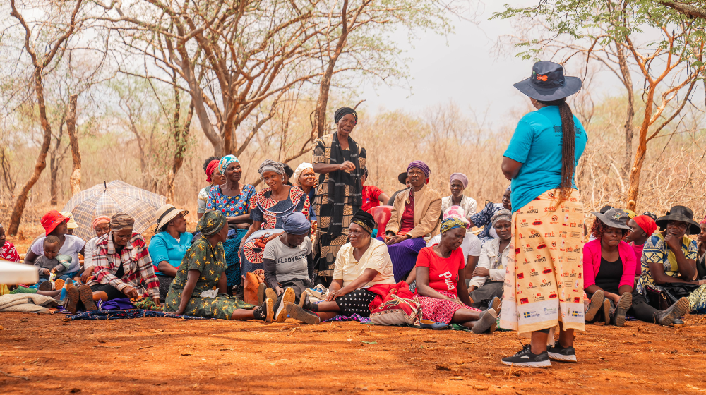 A woman in a blue t-shirt and a sunhat with the UNFPA logo stands before a crowd of women sitting and standing under trees