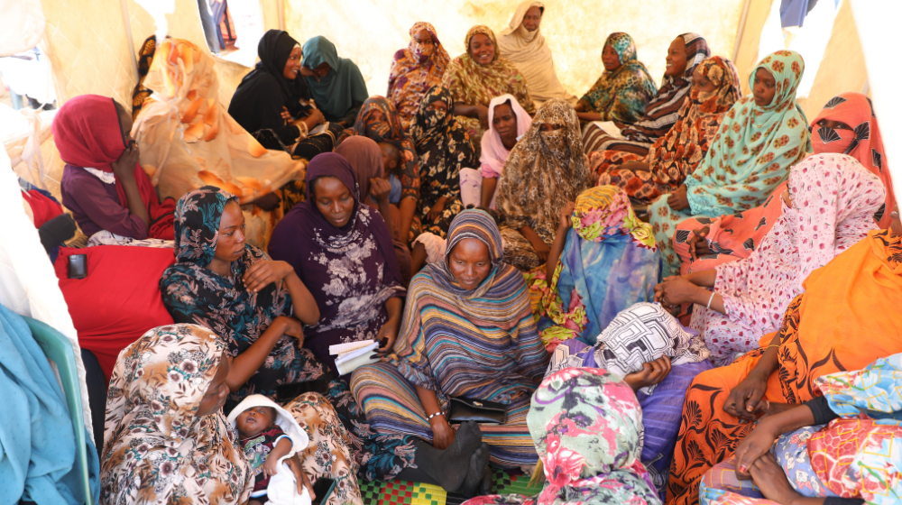 A group of women in colourful, patterned clothes, some holding babies, sit in a circle inside a white-walled tent