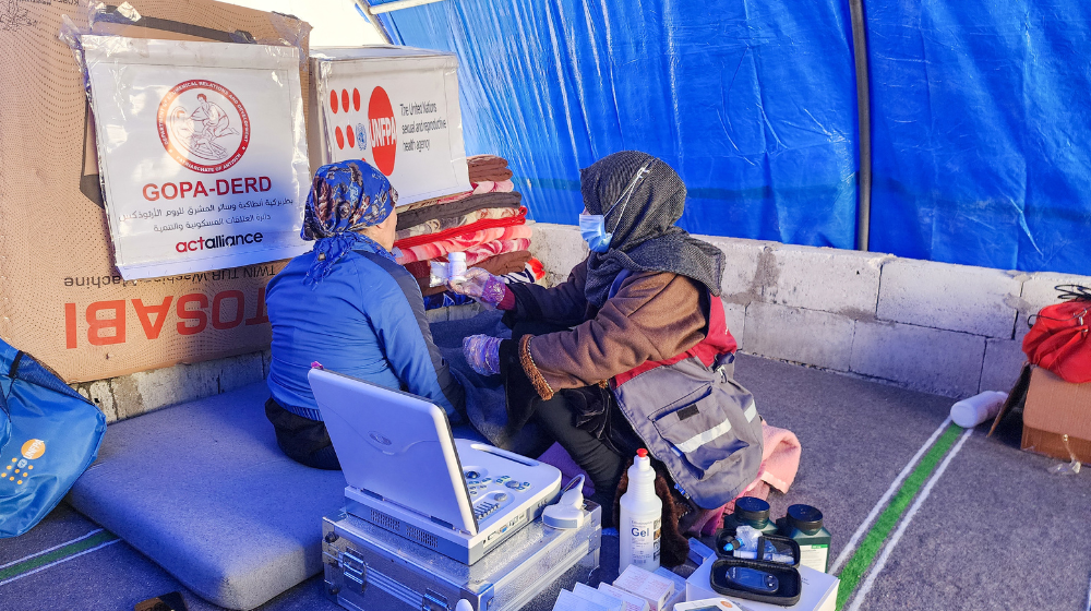  Inside a blue tarpaulin tent, a female health worker is seen from the side sitting in front of a female patient who is on a mattress. Beside them are medical equipment, stacked blankets and boxes with the UNFPA logo