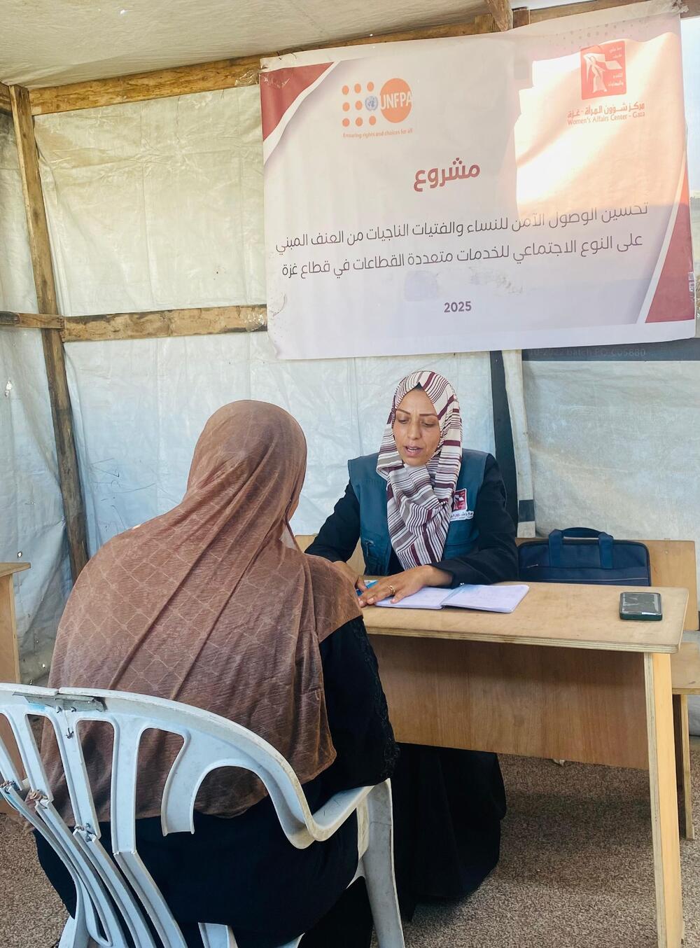 Two women sit facing each other across a wooden desk set up inside a white tarpaulin tent with a gravel floor