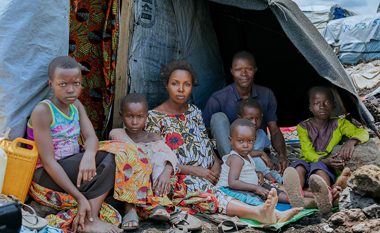  pregnant woman, her husband, and their five children sit together in the shade of a blanket stretched over their makeshift shelter.