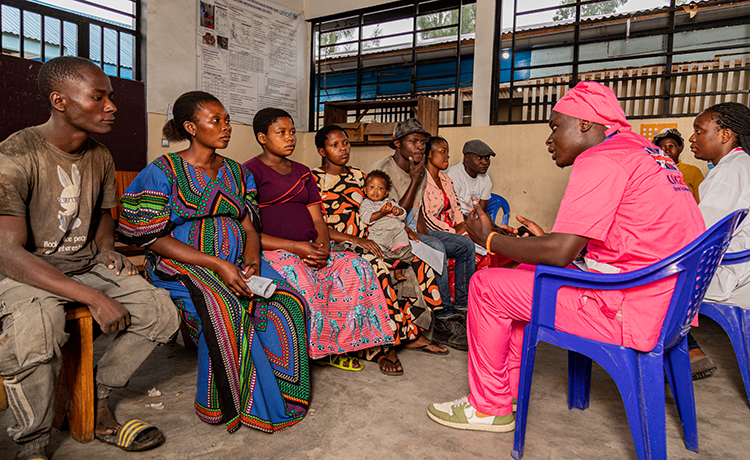  Samuel and Esther sit among seven other adults in a clinic room. A health worker in pink scrubs speaks with them. The UK AID logo is visible on the back of the health worker’s shirt.