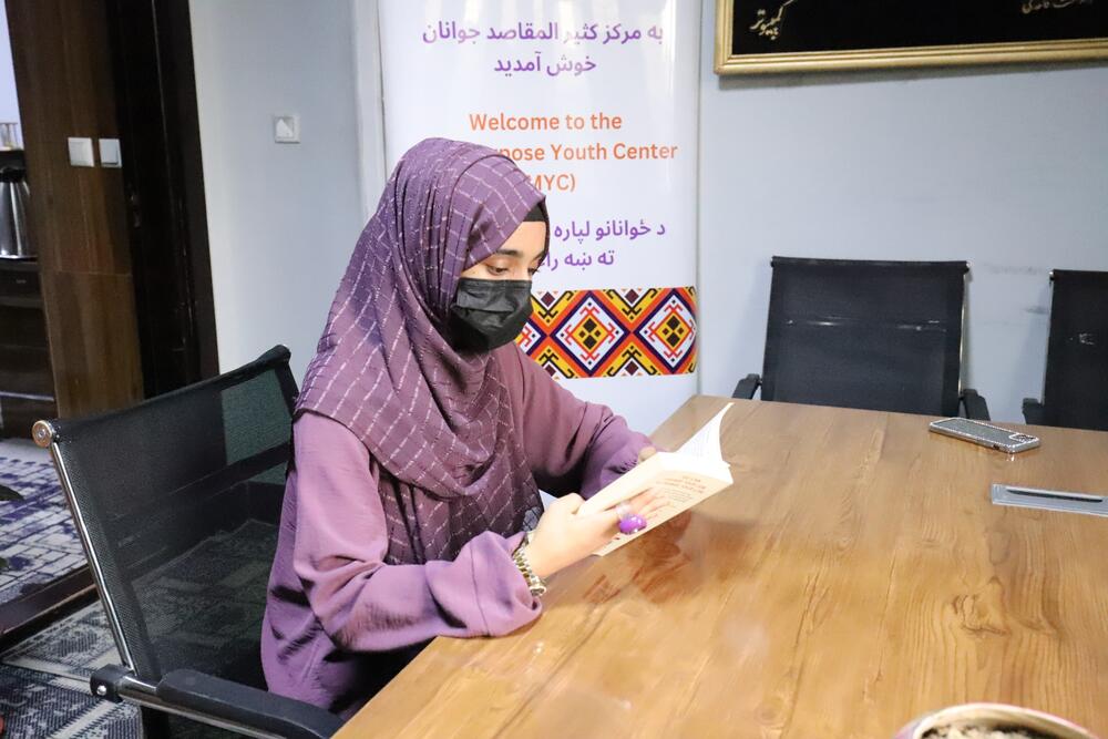 A young woman in purple clothing and headscarf and wearing a black face mask sits at a wooden desk reading a book