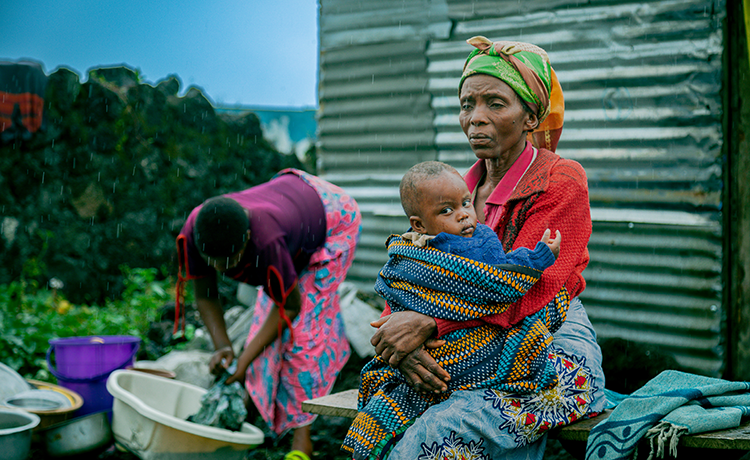  An elderly woman holds her infant grandson outside a shelter of corrugated metal. Behind her, a woman washes clothes in a plastic tub.