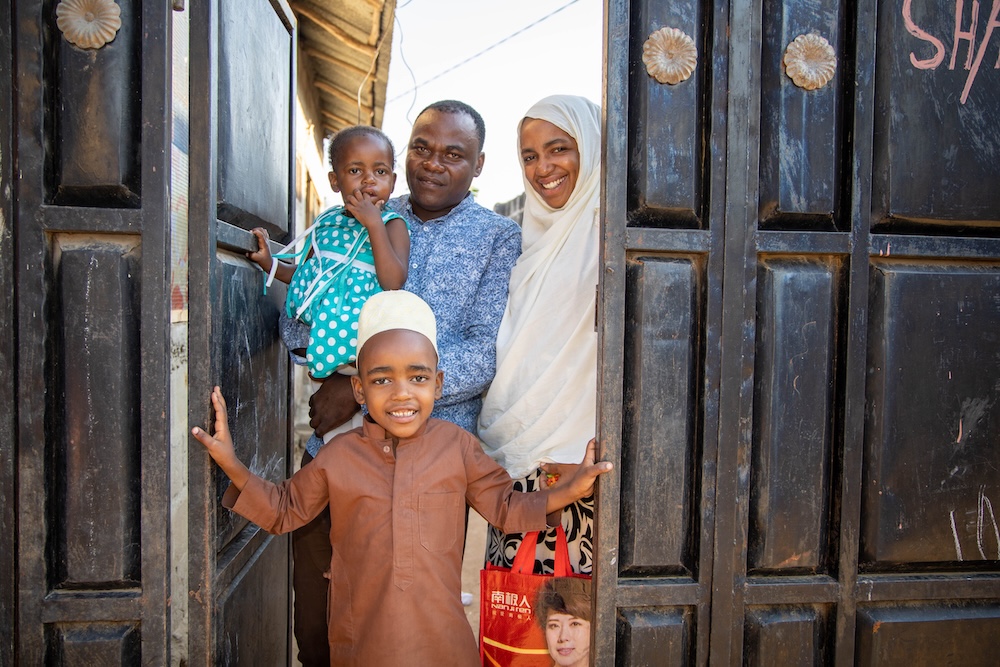  A family of four stands in the doorway of their home. The father holds his toddler daughter. The mother, wearing a white hijab, smiles brightly. Their son is in the foreground holding the door open.