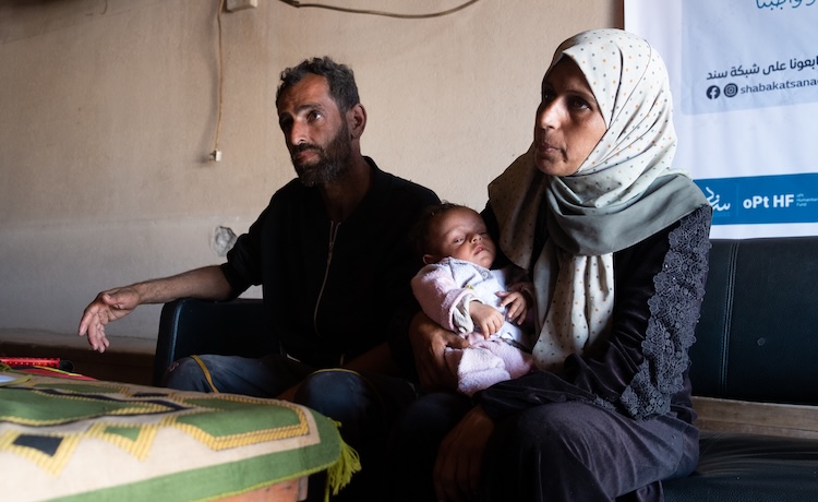 A man and woman sit on a wooden bench. The woman holds a newborn baby.