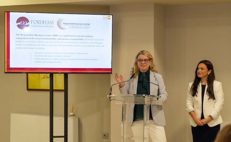 A woman standing at a podium is speaking. To her right is a presentation on a large screen and on her left is another woman. 