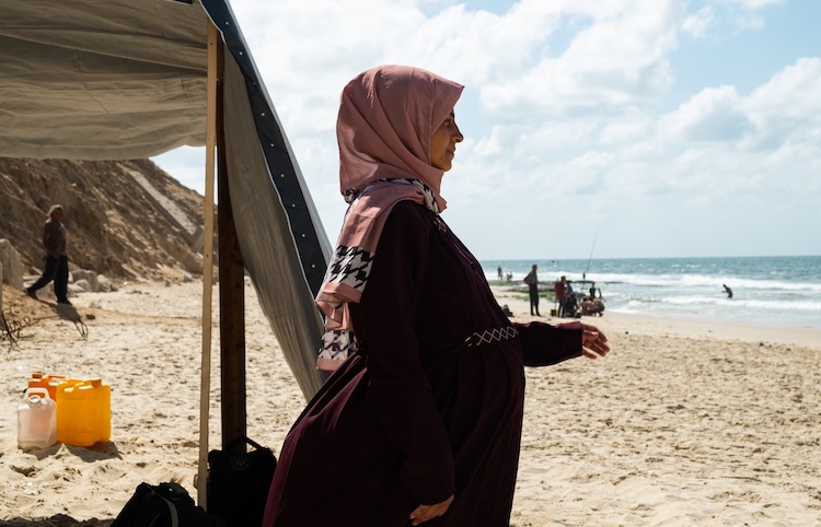 In a pink hijab stands on a beach beside a makeshift tent of tarpaulin and sticks.