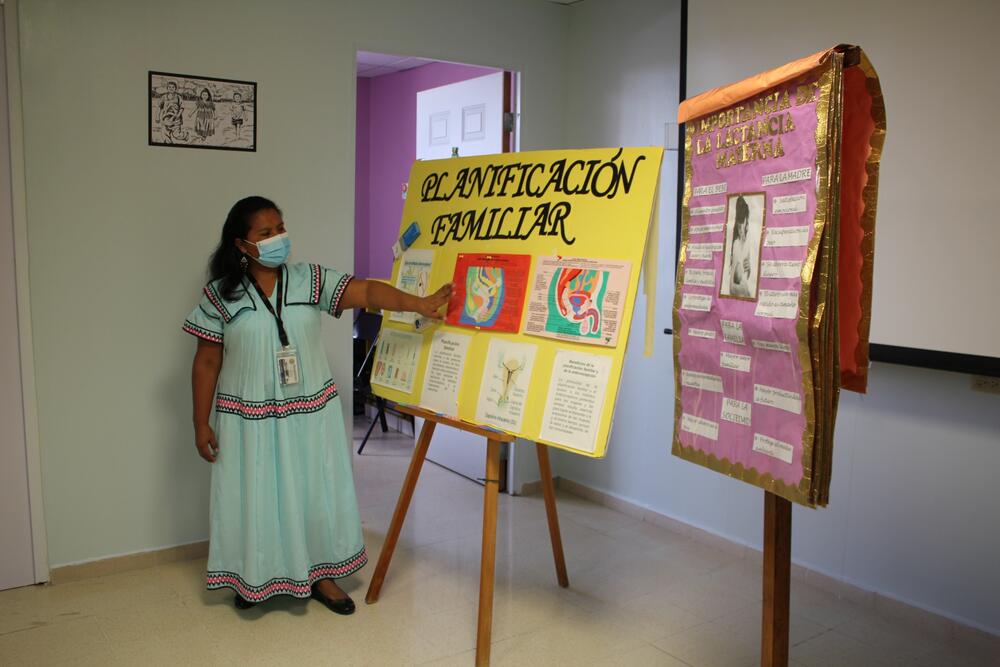 a woman in a turquoise dress and a face mask stands beside a yellow board with “family planning” printed in large letters in Spanish, followed by a series of images and instructions. Bride that is another flipboard in pink with white boxes and gold lettering
