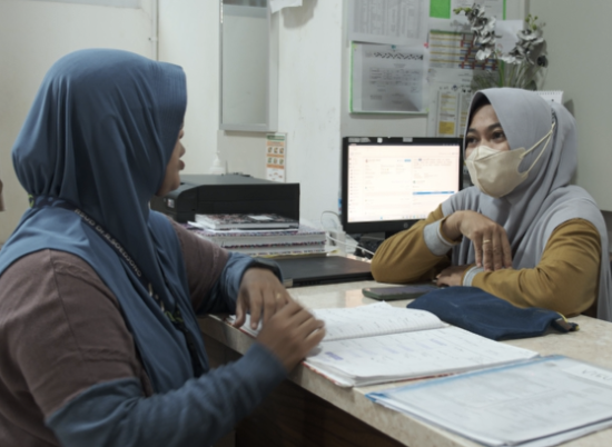 Two women in head coverings sit across from one another in a medical office with a computer in the background and files open in front of them