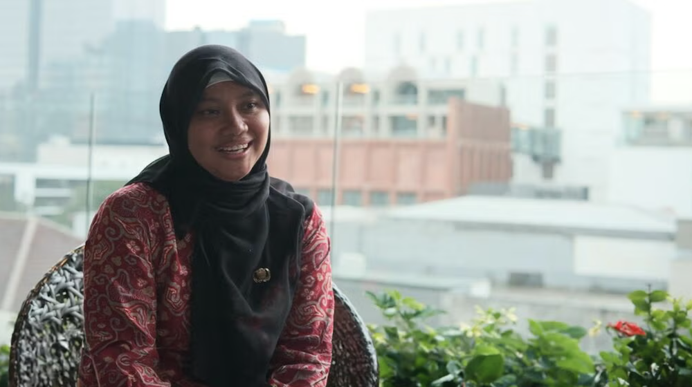 The photo shows Siti Muflihah sitting in front of a window with a view of an urban area.