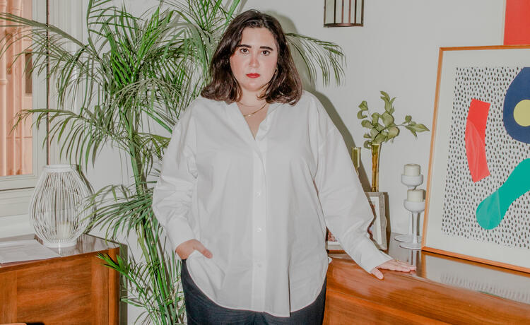 A woman in a white button-down shirt stands in front of an abstract painting and a houseplant. Her face is serious.