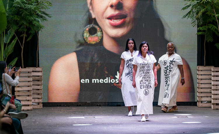  صورة امرأة على شاشة كبيرة فوق مسرح عرض أزياء. ثلاث نساء يقفن على مسرح العرض يرتدين فساتين بيضاء طويلة تحمل شعار "حق الجسد"