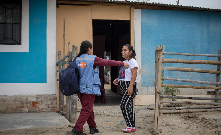A woman in a blue vest with the UNFPA logo reaches out to hug a visibly pregnant woman, who is smiling, in the sandy courtyard of a rural home.