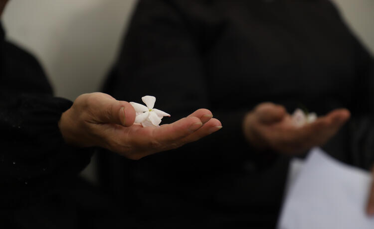 A close-up photo shows a woman’s hand holding a delicate white flower. In the background is another woman also holding a white flower.]