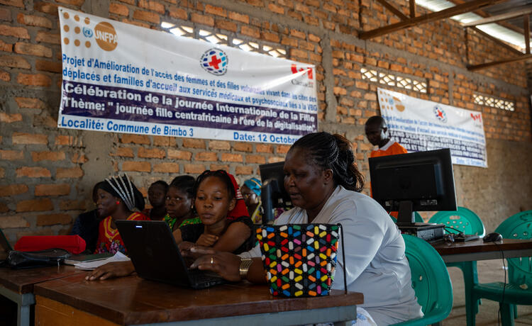 Women and girls sit at desks in a brick-walled room. On the desks are laptop computers and monitor screens. On the wall is a sign with the UNFPA logo, as well as other logos.