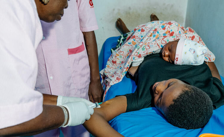Two health workers in pale pink scrubs attend to a young woman lying on a hospital bed as she holds a baby to her chest