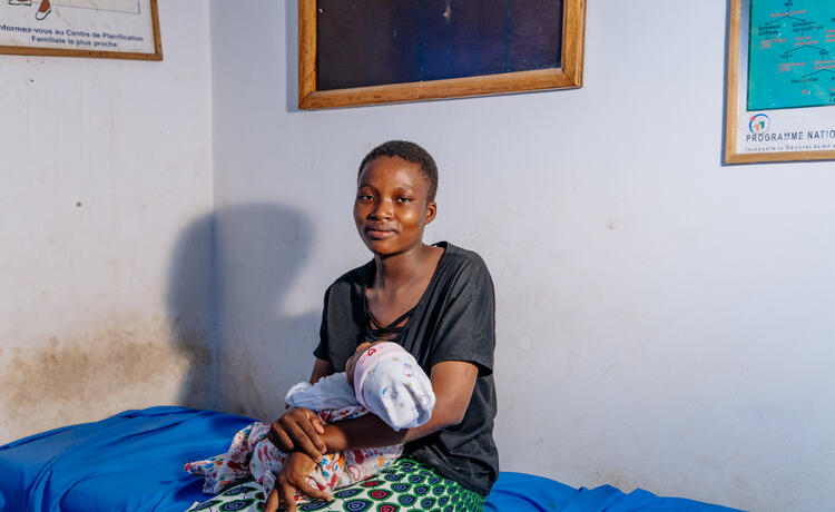 A young woman in a dark grey top sits on the side of a hospital bed holding a baby wearing a bonnet