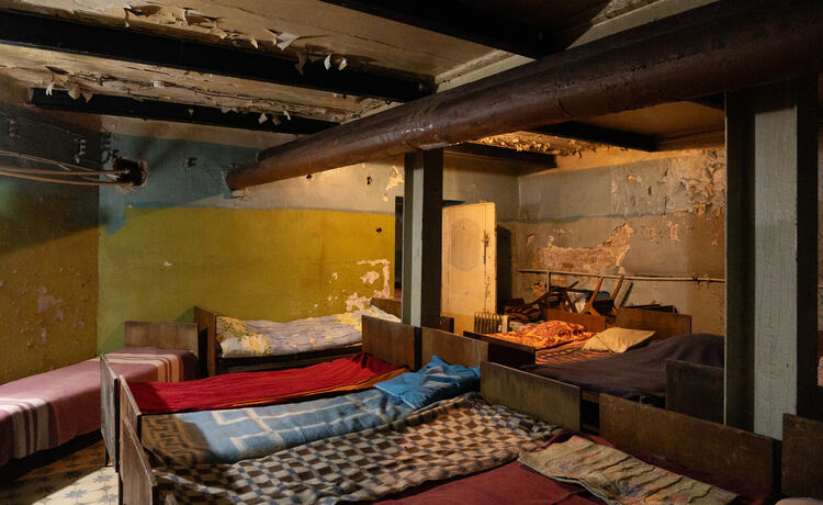 Several single wooden beds with simple sheets on them are crammed together in a bunker, with peeling walls and stacked-up chairs visible in the background