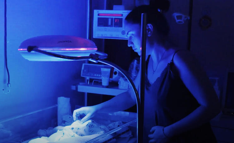 A woman stands beside a baby in an an incubator, bathed in blue light