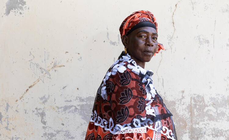 A woman in a red patterned dress and headscarf looks solemnly at the camera.