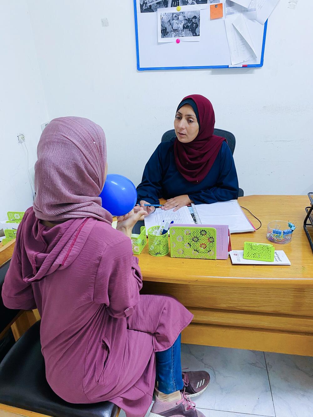 A woman in a dark red headscarf takes notes as she speaks with another woman seated across the desk from her in an office