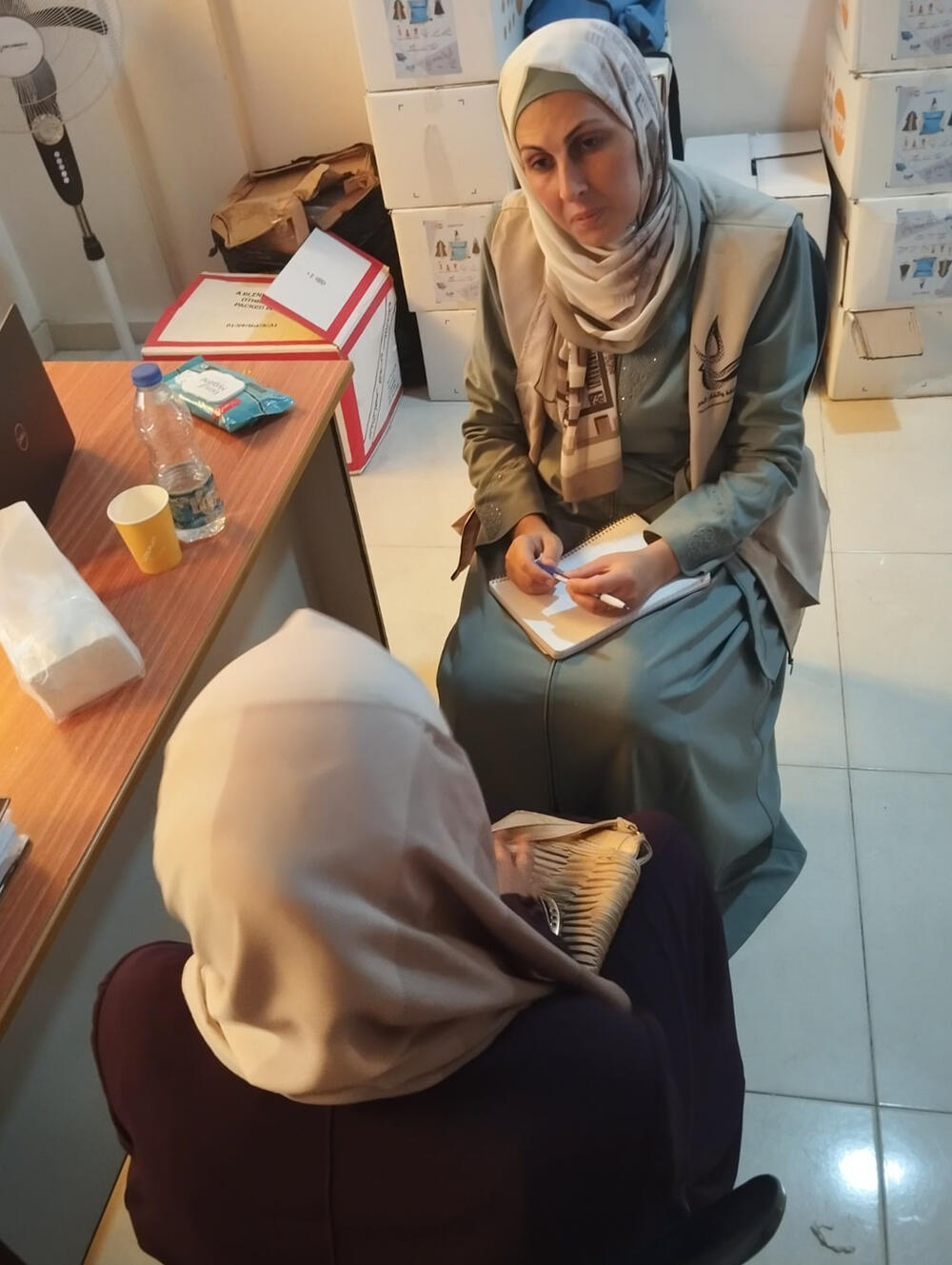  A woman seated in front of a desk in a small office space, dressed in a white headscarf and long green dress, hands a piece of paper to  another woman in a white headscarf, only seen from behind