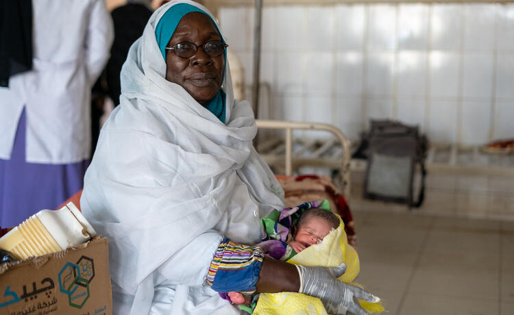 A midwife in a white uniform, blue headscarf and glasses holds a yawning newborn baby on a hospital bed in a maternity ward. The baby is wrapped in a colorful blanket.