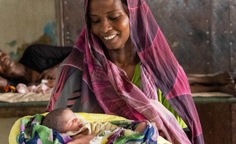 A woman in a pink and purple headscarf cradles her newborn baby. She is smiling widely.