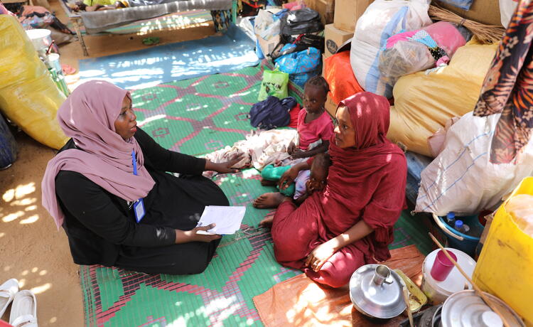 A woman in red sits beside two of her children on a colourful blanket over sand. She speaks to a woman wearing a headscarf and a blue badge.