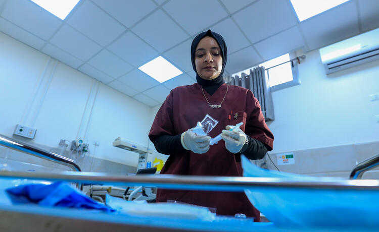 A female health worker in dark red scrubs, white plastic gloves and a black head covering holds medical equipment while standing in a hospital ward.