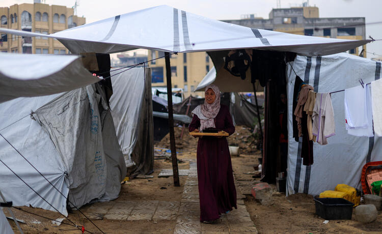 A woman in a long maroon dress and pale flowery head scarf carries a wooden tray as she walks between makeshift tents surrounded by muddy ground.