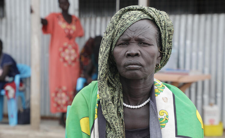 A woman in a dark top, white beaded necklace and patterned green, black and white headscarf and shawl looks into the camera with a serious expression. In the background another woman wearing a bright, long, coral-coloured dress stands near a corrugated metal wall.