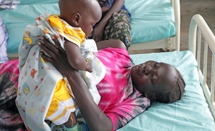 A woman lying on a light blue hospital bed smiles and holds a baby above her. The woman is wearing a pink and black tie-dye top and the baby is wearing a yellow and white outfit. In the background other hospital beds with light blue sheets are visible.