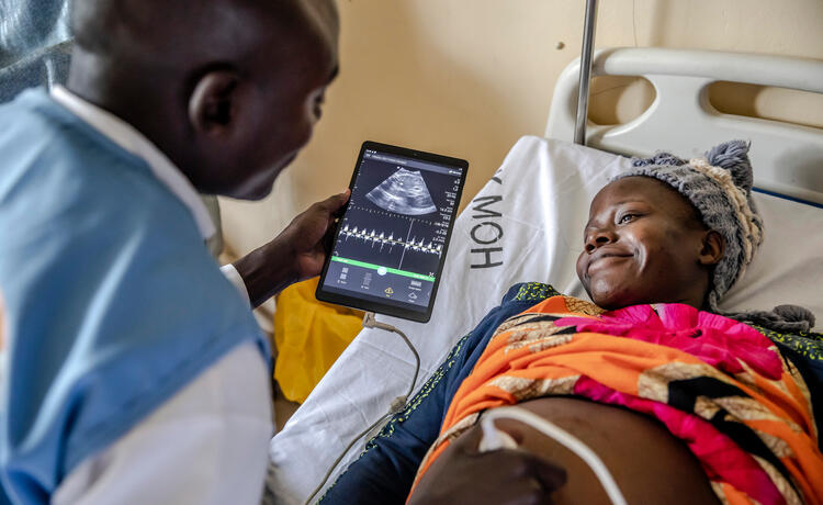 A man in a blue vest holds a tablet that shows an ultrasound image. He is showing it to a pregnant woman, who is smiling brightly.