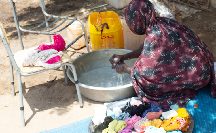 A woman in a blue and purple robe washes clothes by hand in a large tin basin