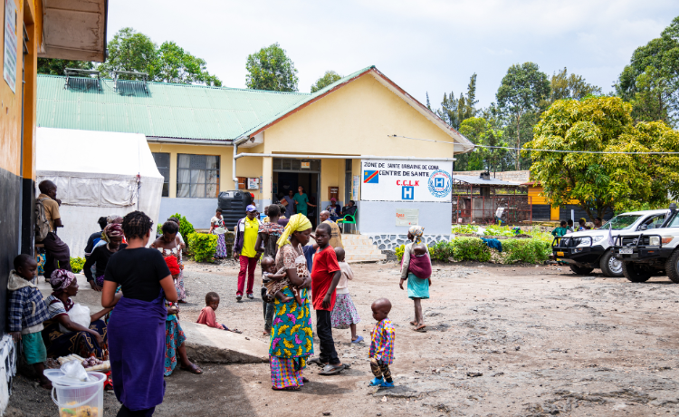 A health clinic is pictured with two cars outside and a number of people waiting in the foreground