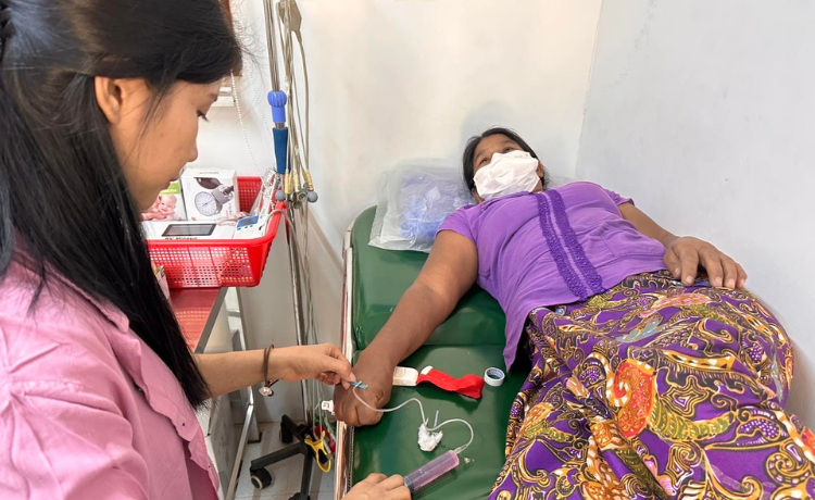  A health worker in a pink coat administers medicine to a woman lying on a hospital bed, wearing a white facemask, purple shirt and patterned skirt]