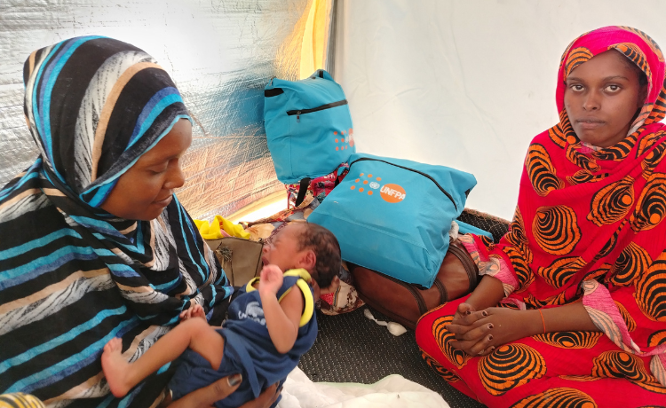 A woman in a striped headscarf holds a newborn baby; she is sitting on the floor of a tent beside another woman and dignity kits displaying the UNFPA logo.