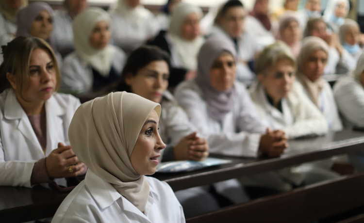  A midwife in a white coat and headscarf speaks in the foreground; behind her are many other midwives in white coats.
