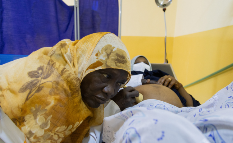 A midwife in a yellow headscarf listens to a pregnant woman’s belly