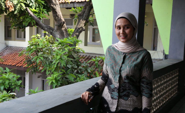 A woman in a green and white patterned outfit and white head covering smiles on a balcony