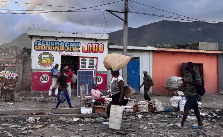  People walk along a dirt road, carrying packages and buckets, in front of a shop and with mountains visible in the background